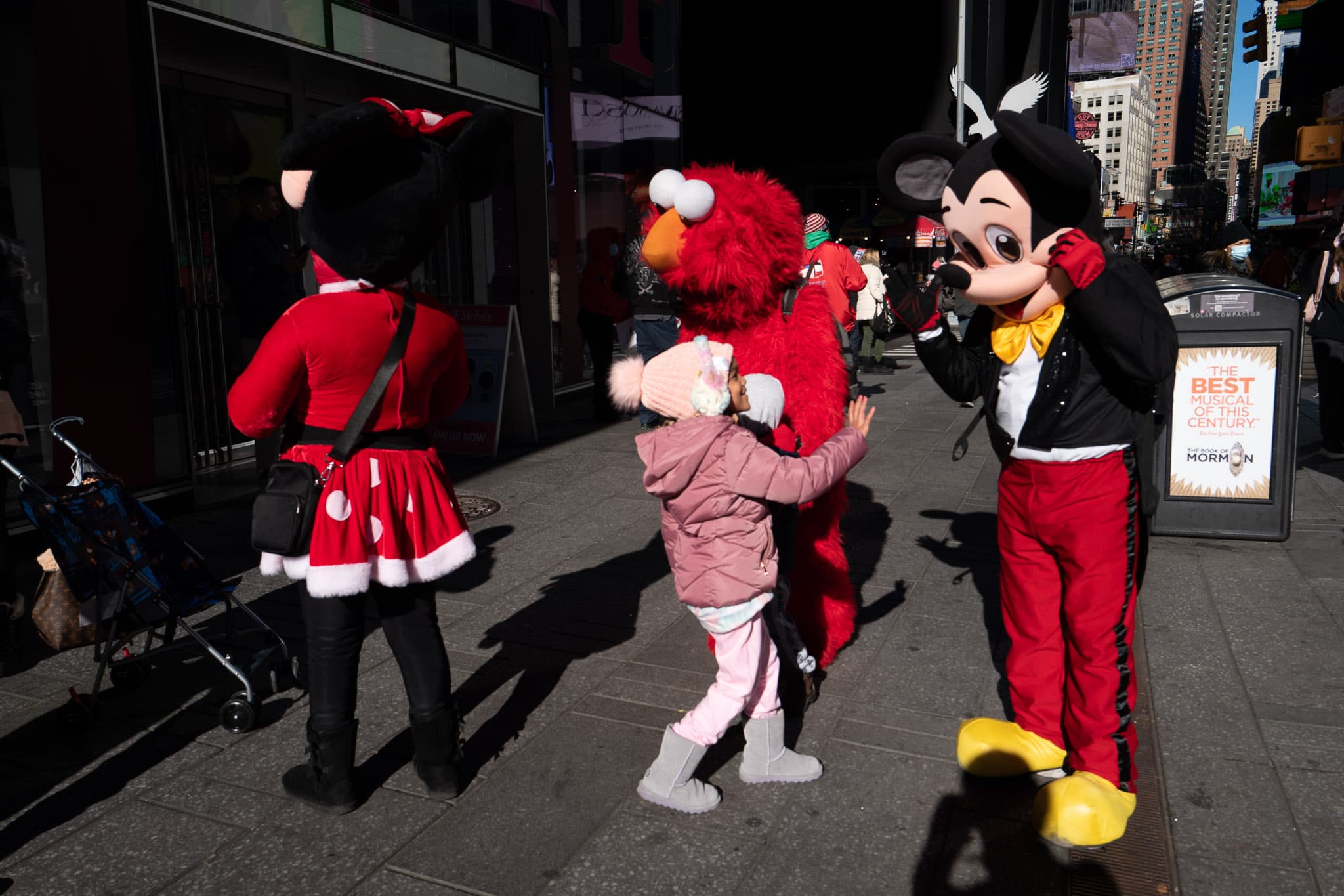 A child waves hello to a Mickey Mouse character in Times Square of New York City. Elmo and Minnie Mouse can be seen in the background. 