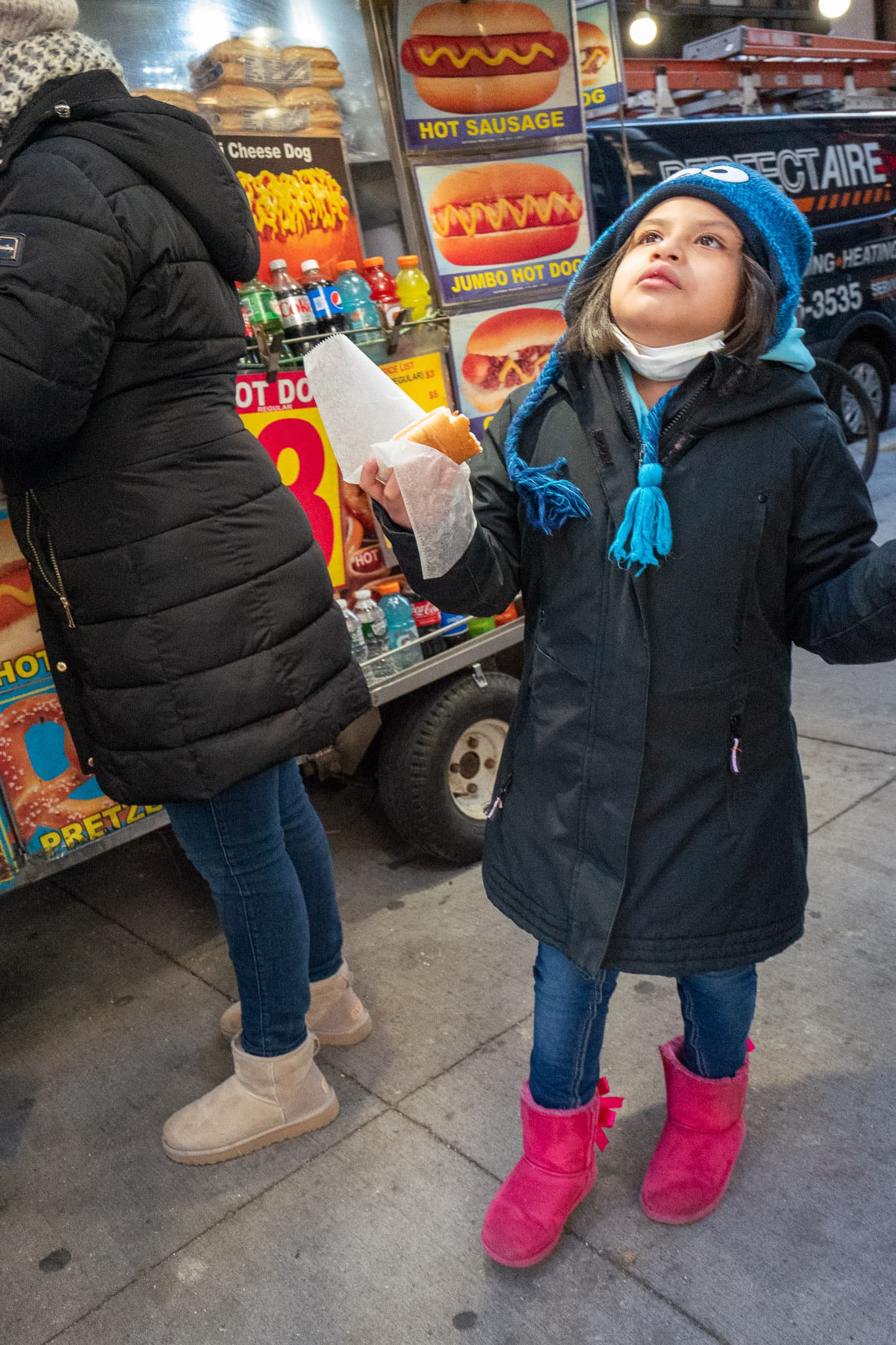 A child looks up at the sky while enjoying a hot dog purchased from the hot dog stand in the background.